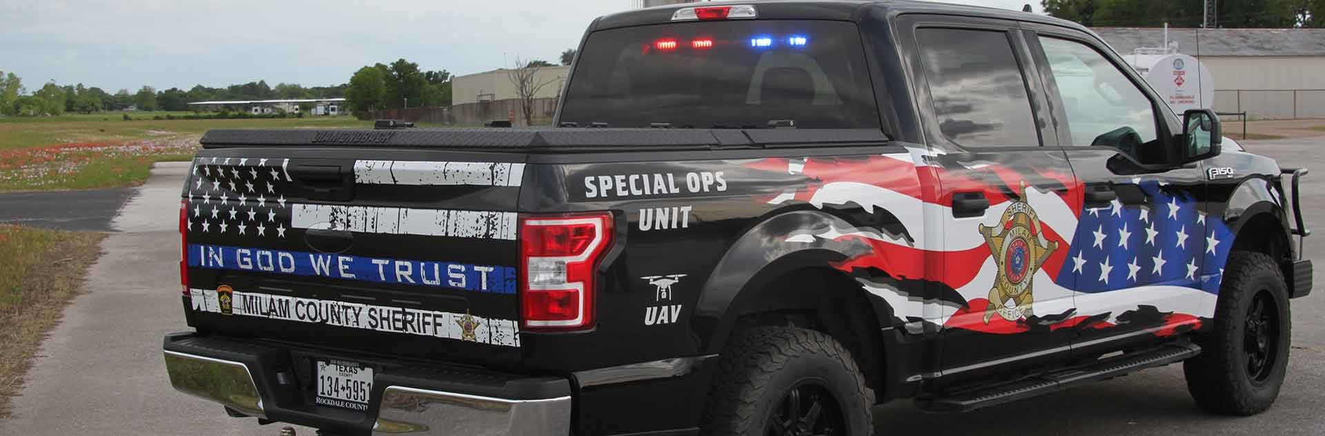 Black 4-door extended cab truck with red and blue lights in the back window. On the tail gate a black and white American flag, In God We Trust in white text, Milam County Sheriff in black text, Special Ops Unit in white text on the passenger side back top fender, and UAV with an outline of a drone in white text on the passenger side back bottom fender. On the passenger side back door is the Milam County Sheriff’s Office badge. Across the front and back passenger side doors is a partial American Flag.